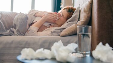 Foto van vrouw die met griep ziek in bed ligt en haar neus snuit. Op de voorgrond zakdoekjes