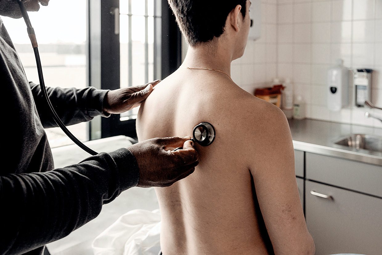 Afbeelding van huisarts die met een stethoscoop een patient op de rug luistert.